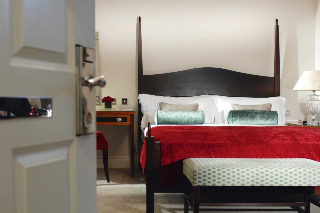 Macdonald Bath Spa Hotel Rom bilde The photo shows a well-decorated hotel room. In the foreground, there is a partially opened door leading into the room. The focal point of the room is a large bed with a dark wooden headboard and stylish red and white bedding, accented with green pil