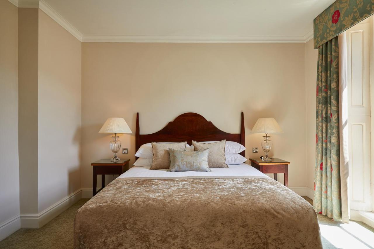 Macdonald Bath Spa Hotel Eksteriør bilde The photo shows a cozy hotel bedroom. It features a large bed with a plush, light-colored bedspread and several pillows. On either side of the bed, there are wooden nightstands with lamps providing soft lighting. The walls are painted a neutral color
