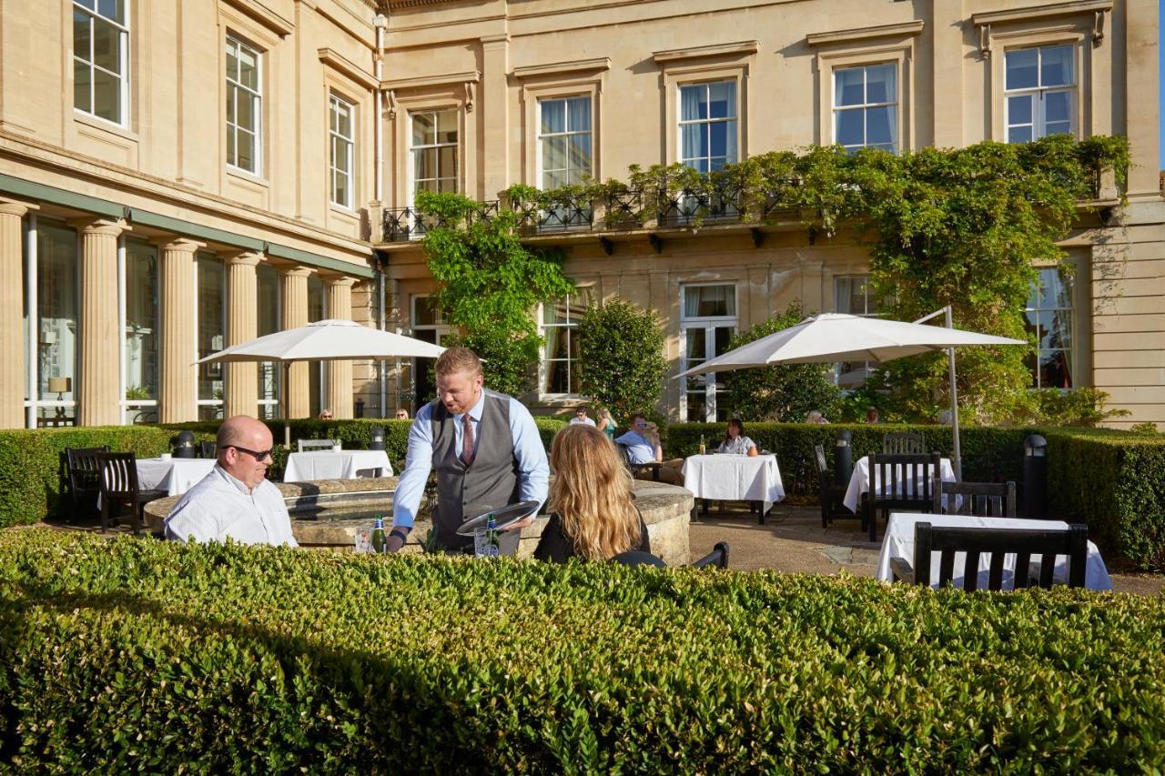 Macdonald Bath Spa Hotel Eksteriør bilde The photo depicts an outdoor dining area of a restaurant. In the foreground, there are neatly trimmed hedges surrounding the dining space. Two patrons are seated at a table; one is a man wearing sunglasses and the other is a woman facing him. A waite