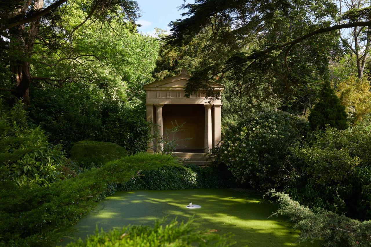 Macdonald Bath Spa Hotel Eksteriør bilde The photo shows a serene garden scene featuring lush greenery and a tranquil pond. In the background, there is a classical-looking structure with columns, resembling a small temple or pavilion. The pond appears to have vegetation covering its surface
