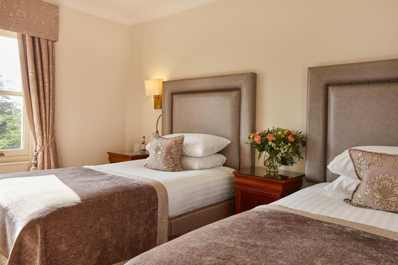 Macdonald Bath Spa Hotel Eksteriør bilde The photo shows a hotel room featuring two beds with elegant headboards. Each bed is neatly made with white linens and a soft, decorative throw blanket. There are matching pillows on the beds, and a small, stylish bouquet of flowers is placed on a wo