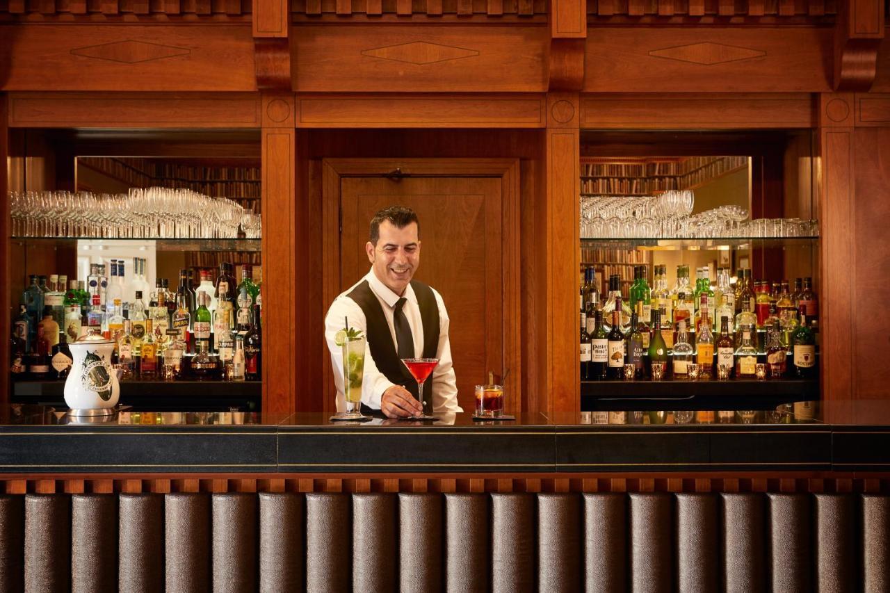 Macdonald Bath Spa Hotel Eksteriør bilde The photo depicts a bartender standing behind a polished bar. He is wearing a formal outfit with a vest and a bow tie, and he appears to be smiling as he presents a cocktail. Behind him is a well-stocked bar shelf filled with various bottles of liquo