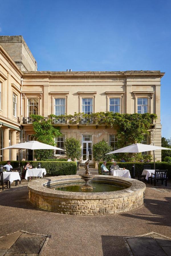 Macdonald Bath Spa Hotel Eksteriør bilde The photo depicts a charming outdoor dining area of a classic-looking building with elegant architecture. The structure features large windows and is adorned with greenery, including climbing plants. In the foreground, there is a circular fountain, a
