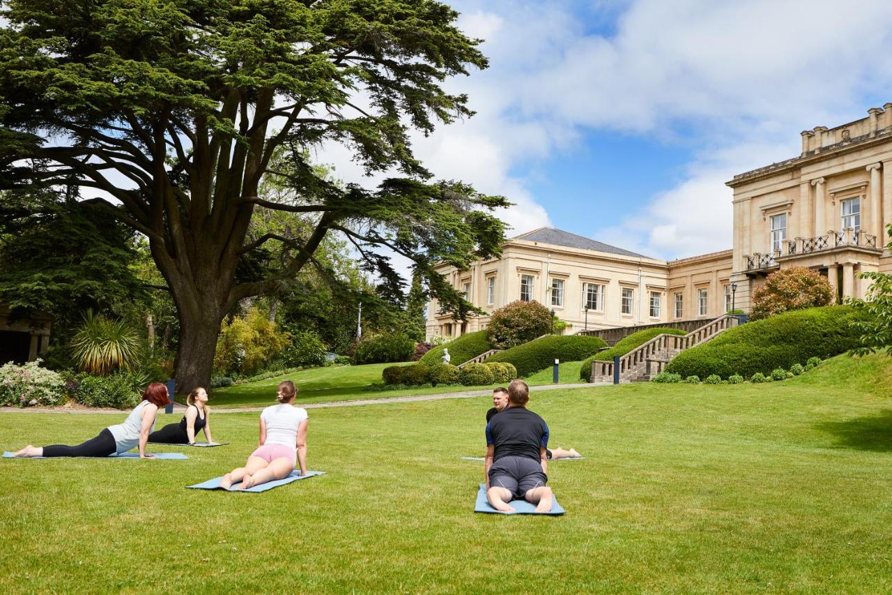 Macdonald Bath Spa Hotel Eksteriør bilde The photo shows a group of people engaging in a yoga session outdoors. They are positioned on yoga mats on a lush green lawn in front of a large, elegant building with columns. Behind them, there are trees and well-maintained gardens, contributing to