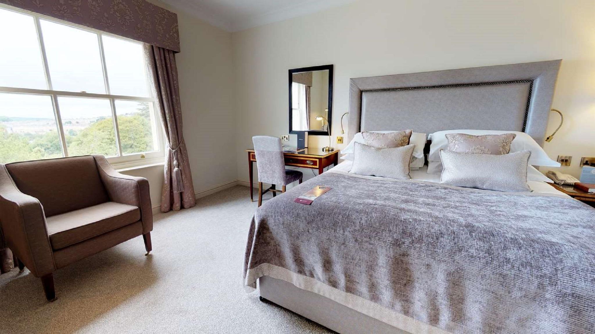 Macdonald Bath Spa Hotel Eksteriør bilde The photo shows a cozy and well-decorated bedroom. It features a large bed with a plush, grey bedspread and several decorative pillows. Beside the bed, there is a wooden bedside table with a lamp. A small desk and chair are positioned near a window, 