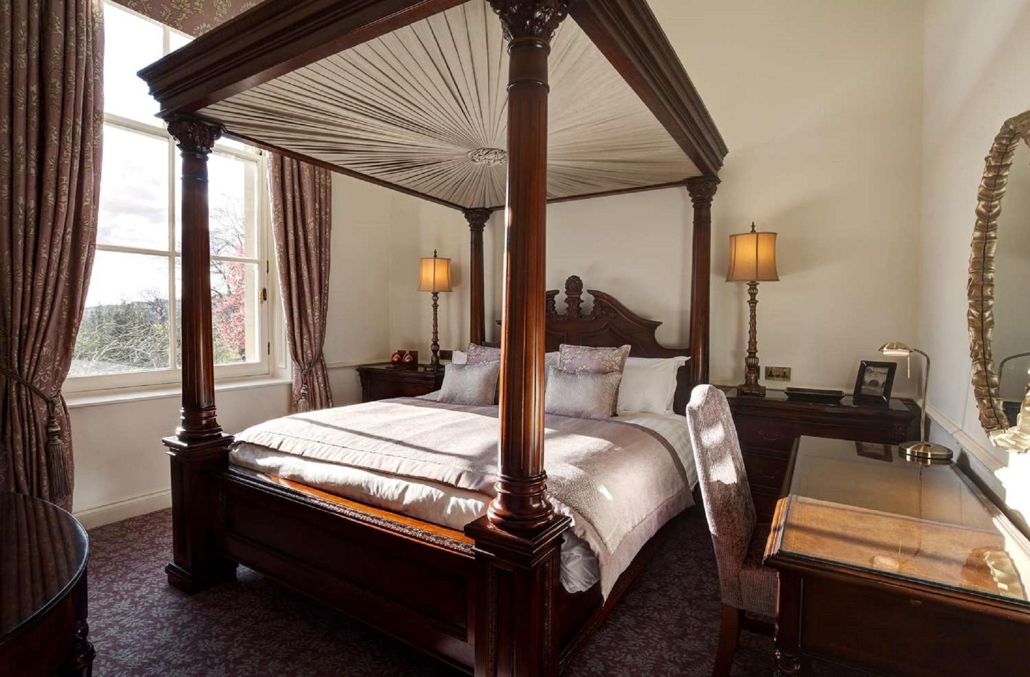 Macdonald Bath Spa Hotel Eksteriør bilde The photo shows a well-decorated bedroom featuring a four-poster bed with an elegant wooden frame. The bed is dressed with light-colored linens and several decorative pillows. On either side of the bed, there are matching bedside lamps that provide s