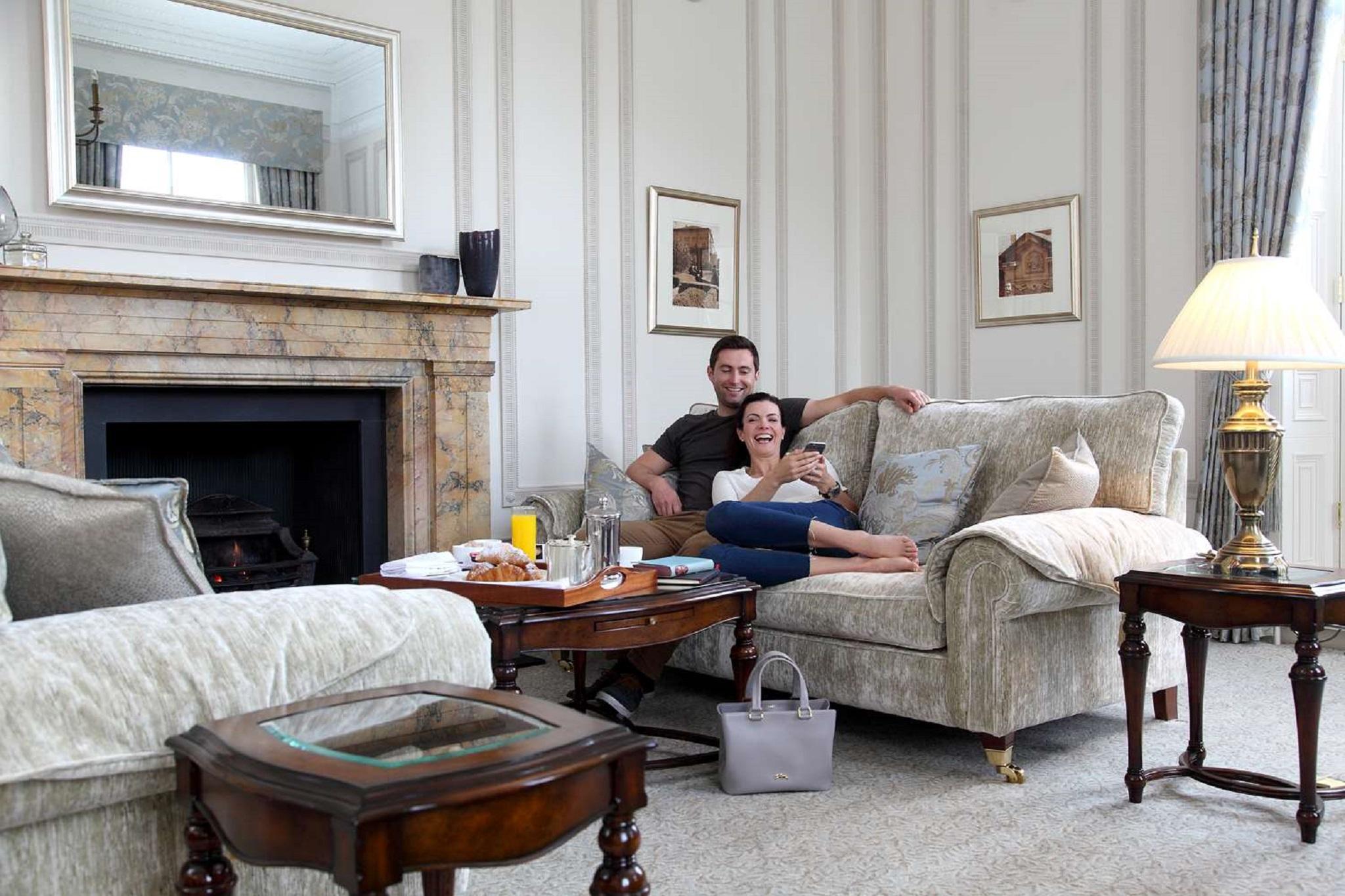Macdonald Bath Spa Hotel Eksteriør bilde The photo shows a cozy living room setting where a couple is sitting together on a light-colored sofa. They appear relaxed, with the man sitting slightly behind the woman, who is smiling and looking at her phone. The room features elegant decor, incl