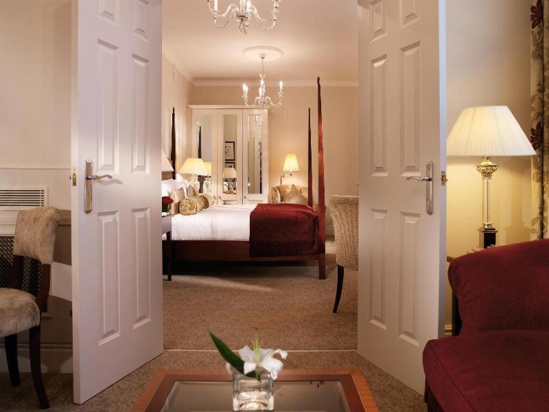 Macdonald Bath Spa Hotel Rom bilde The photo shows a hotel room that can be viewed through open double doors. Inside, there is a neatly made bed with decorative pillows, a plush area rug, and soft lighting from lamps. The room features elegant furnishings, including a chair, a coffee 